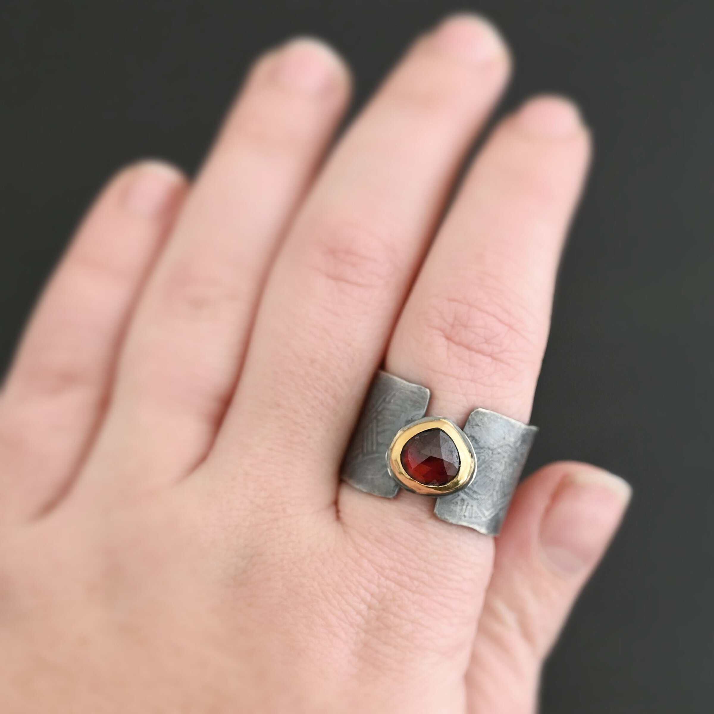 "Fire Of Love" Ring. Made out of sterling silver, gold, and garnet. Pear shaped garnet pointing up in the center of the ring set in gold. The gold sits on a layer of silver. The two sides of the band do not touch in the center. The garnet setting connects them together. The ring band is engraved with lines. Ring is worn on a models middle finger.