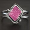 "Bubblegum Cuff." There is a diamond shaped pink druzy in the center. There are two small circular black onyx stones on the bottom right side of the stone. There is silver ball detail around the bottom right and top left of the cuff. There are two bands for the cuff that are thick silver and hammered. The top and bottom band meet together at the back on each side to create the band.