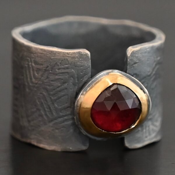"Fire Of Love" Ring. Made out of sterling silver, gold, and garnet. Pear shaped garnet pointing up in the center of the ring set in gold. The gold sits on a layer of silver. The two sides of the band do not touch in the center. The garnet setting connects them together. The ring band is engraved with lines.