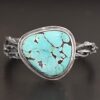 "Under The Big Sky" Cuff. There is a very large, mostly circular turquoise in the center of a thick cuff. The cuff is multiple casted sterling silver twigs that are soldered together and create a cuff.