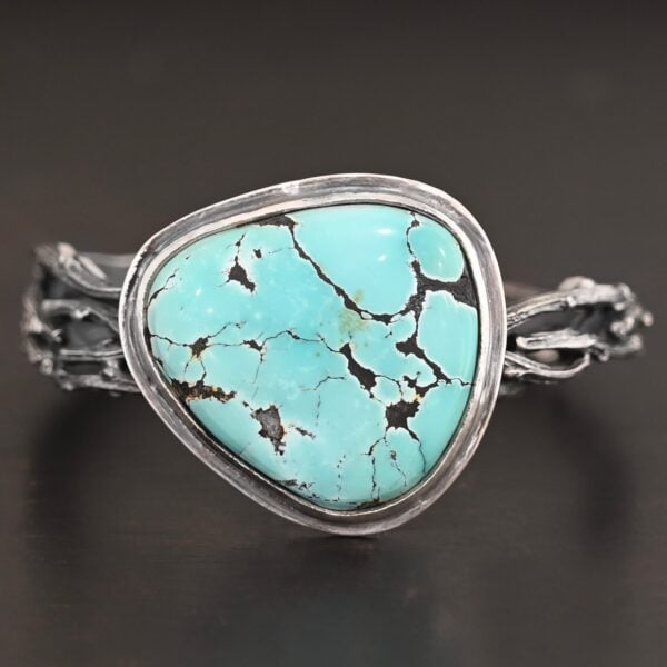 "Under The Big Sky" Cuff. There is a very large, mostly circular turquoise in the center of a thick cuff. The cuff is multiple casted sterling silver twigs that are soldered together and create a cuff.