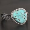 "Under The Big Sky" Cuff. There is a very large, mostly circular turquoise in the center of a thick cuff. The cuff is multiple casted sterling silver twigs that are soldered together and create a cuff.
