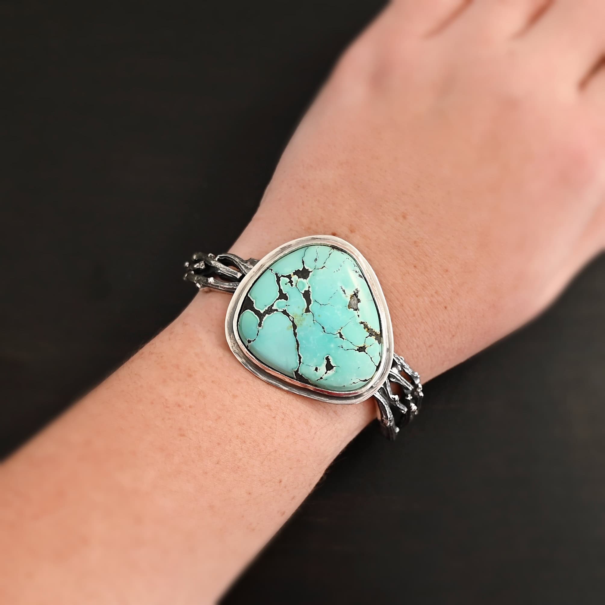Under The Big Sky Cuff "Under The Big Sky" Cuff. There is a very large, mostly circular turquoise in the center of a thick cuff. The cuff is multiple casted sterling silver twigs that are soldered together and create a cuff. Cuff is shown up close by a model.