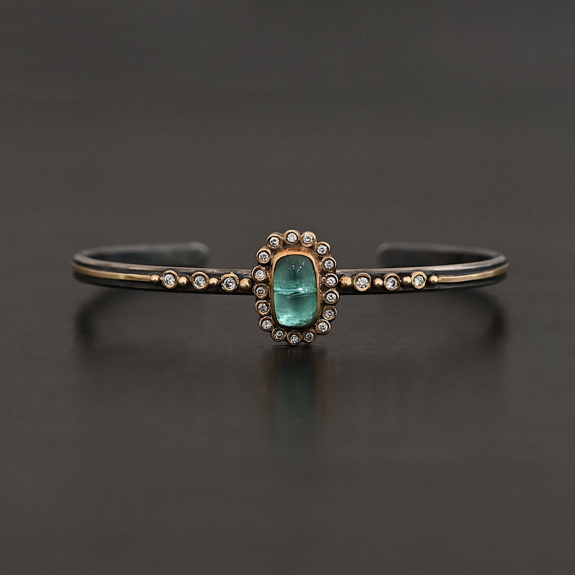 "Secret Of The Sea" Cuff. There is a rectangular, vertical tourmaline in the center, set in gold, with small diamonds surrounding the entire tourmaline. There are seventeen diamonds around the tourmaline. There are six more, three on each side of the cuff. There is gold ball detail between each diamond, and a stripe of gold that goes across the rest of the cuff.