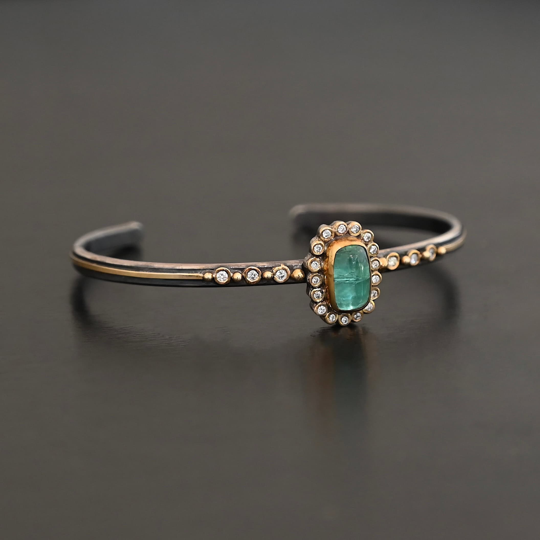 "Secret Of The Sea" Cuff. There is a rectangular, vertical tourmaline in the center, set in gold, with small diamonds surrounding the entire tourmaline. There are seventeen diamonds around the tourmaline. There are six more, three on each side of the cuff. There is gold ball detail between each diamond, and a stripe of gold that goes across the rest of the cuff.