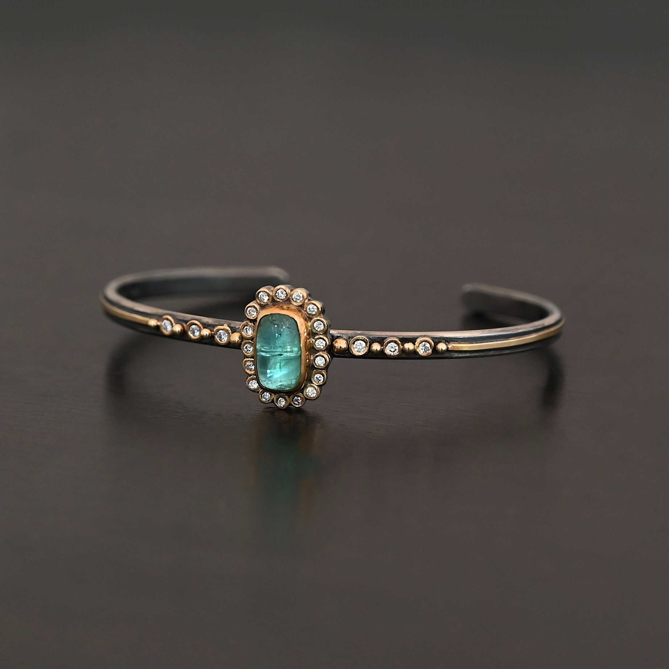 "Secret Of The Sea" Cuff. There is a rectangular, vertical tourmaline in the center, set in gold, with small diamonds surrounding the entire tourmaline. There are seventeen diamonds around the tourmaline. There are six more, three on each side of the cuff. There is gold ball detail between each diamond, and a stripe of gold that goes across the rest of the cuff.