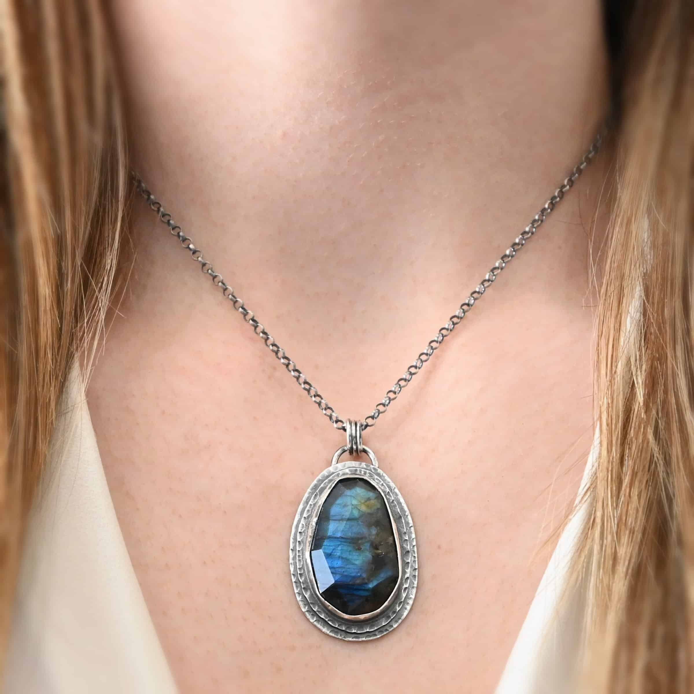A New Day Necklace. There is a large, rose cut, vertical, oval labradorite in the center with two layers of sterling silver around it. There is a loop soldered to the back with three jump rings attached to it that the chain of the necklace goes through. Necklace is shown on a model.