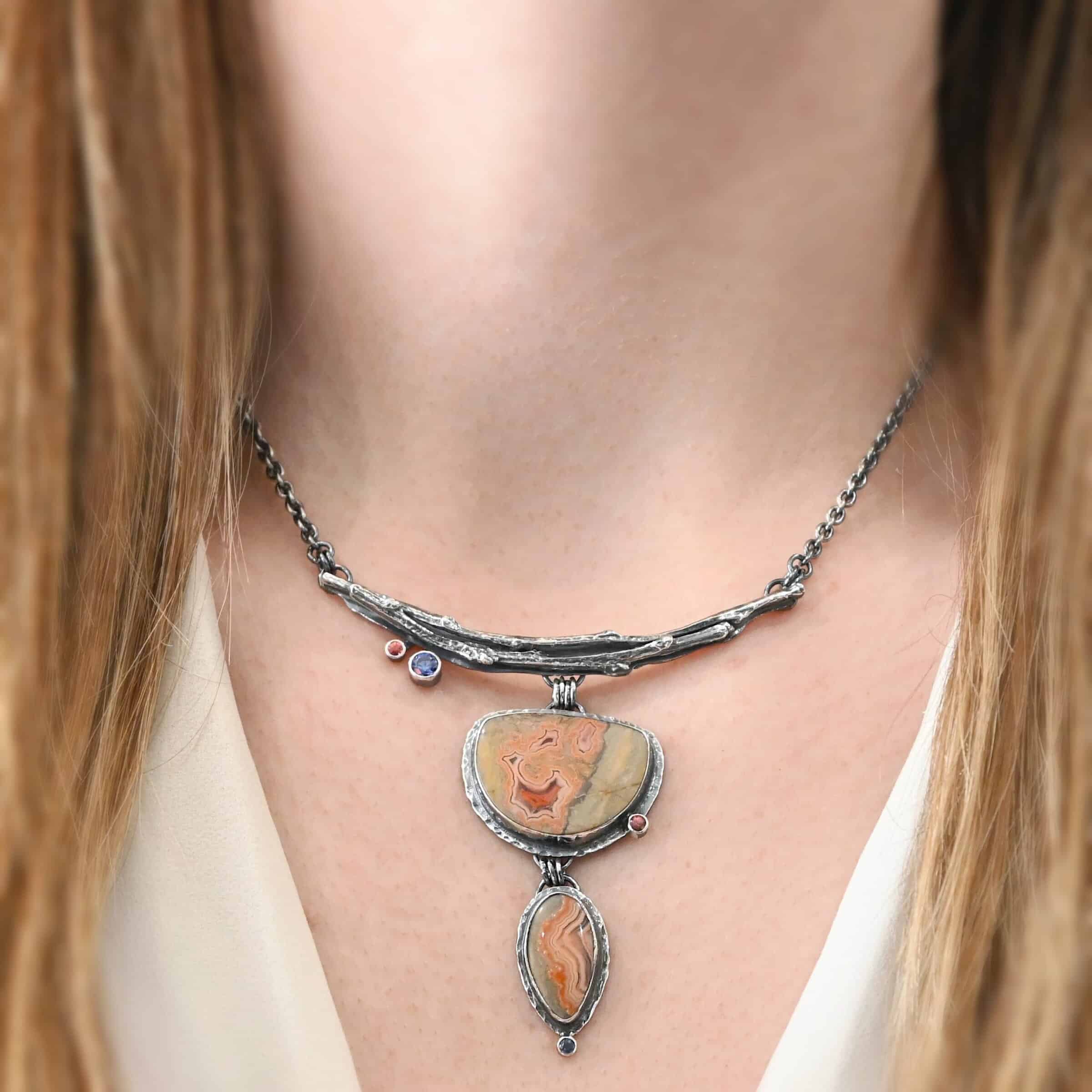 Dare To Be Necklace. There are two curved, casted sterling silver twigs from a branch at the top. On the left side bottom are two small stones. Hanging from the center is a semicircle ocean jasper with a small circular stone on the right side, and beneath that is an oval ocean jasper, vertical. On the bottom of that is a small circular stone. Necklace is shown up close by a model.