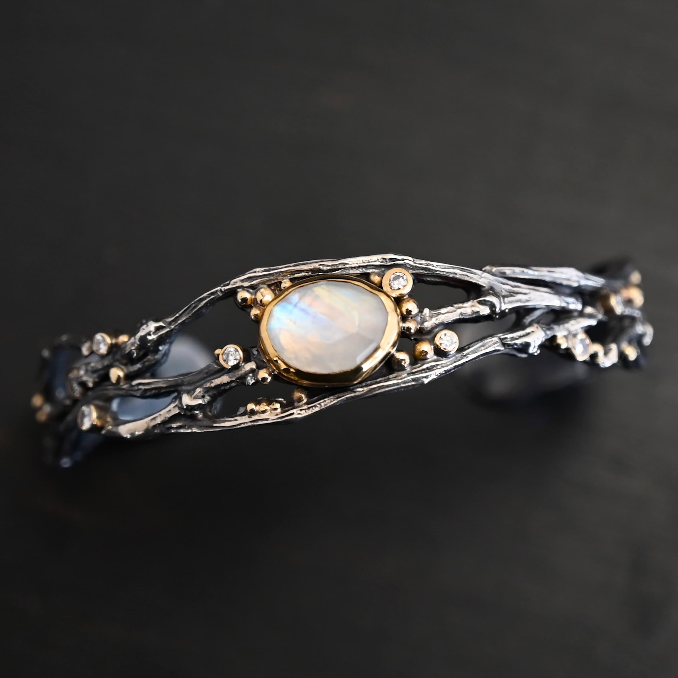 Night Flowers Cuff "Night Flowers" Cuff. There is a moonstone set in gold in the center of a cuff. The band of the cuff is multiple casted sterling silver twigs that intercept. There are small diamonds scattered around it, as well as gold ball detail.