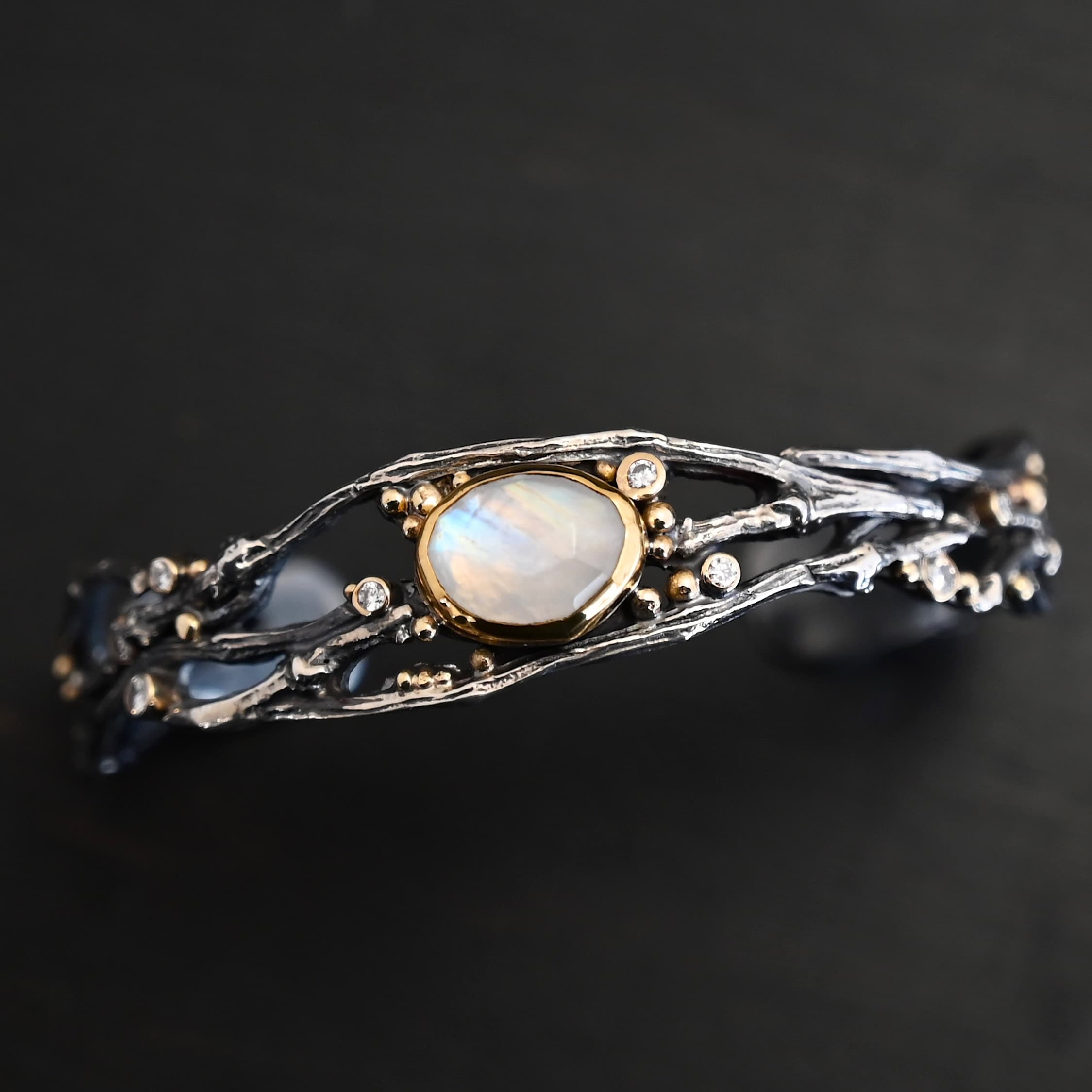 Night Flowers Cuff "Night Flowers" Cuff. There is a moonstone set in gold in the center of a cuff. The band of the cuff is multiple casted sterling silver twigs that intercept. There are small diamonds scattered around it, as well as gold ball detail.