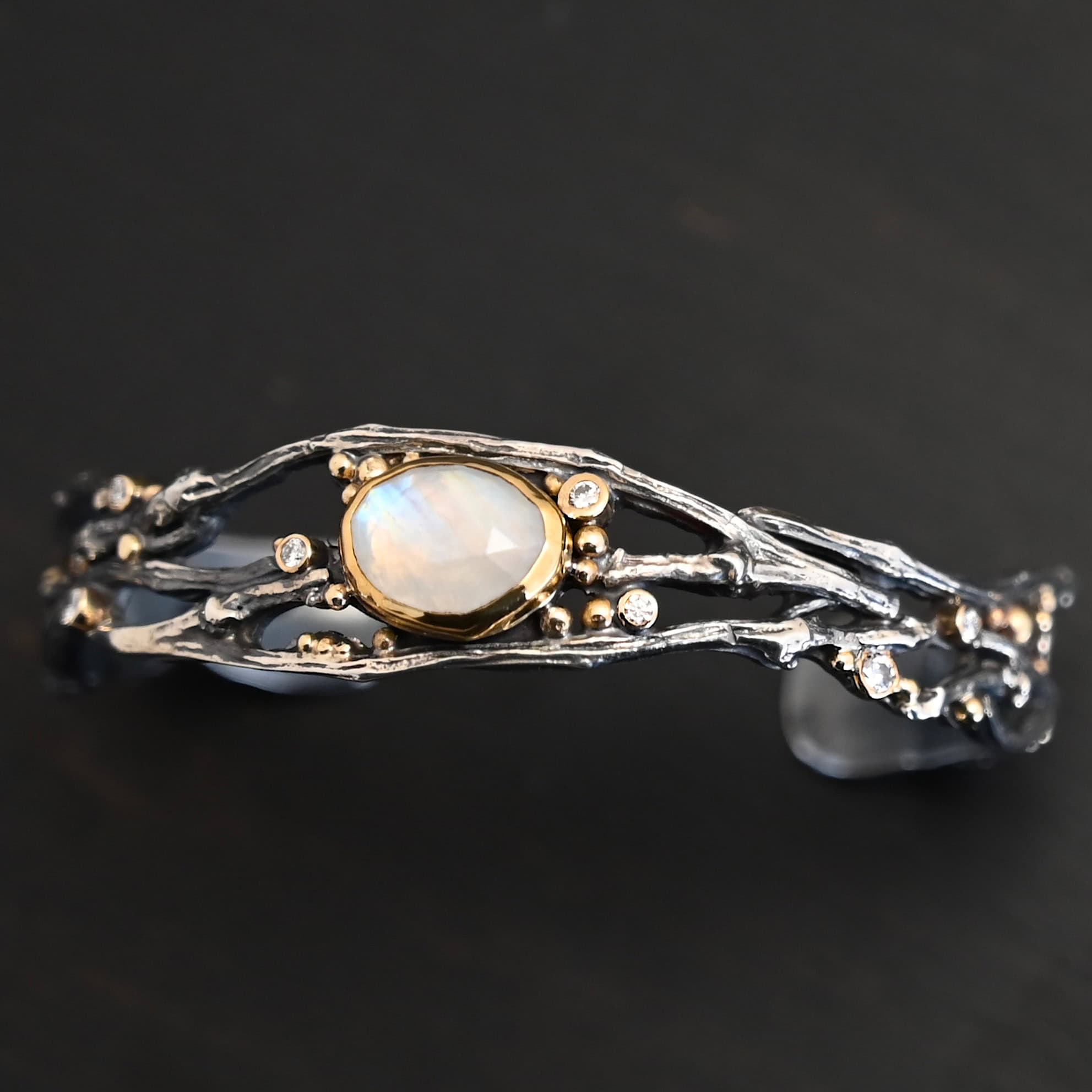 Night Flowers Cuff "Night Flowers" Cuff. There is a moonstone set in gold in the center of a cuff. The band of the cuff is multiple casted sterling silver twigs that intercept. There are small diamonds scattered around it, as well as gold ball detail.