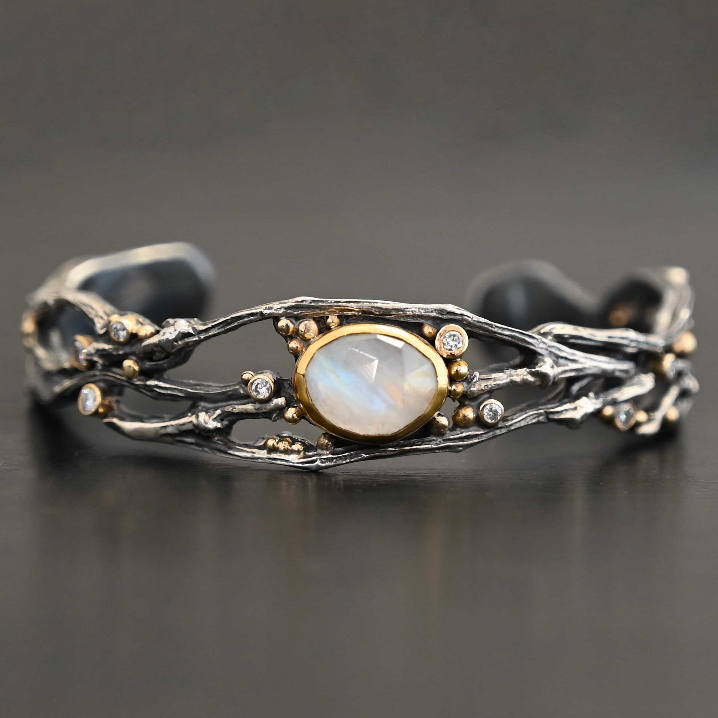 Night Flowers Cuff "Night Flowers" Cuff. There is a moonstone set in gold in the center of a cuff. The band of the cuff is multiple casted sterling silver twigs that intercept. There are small diamonds scattered around it, as well as gold ball detail.
