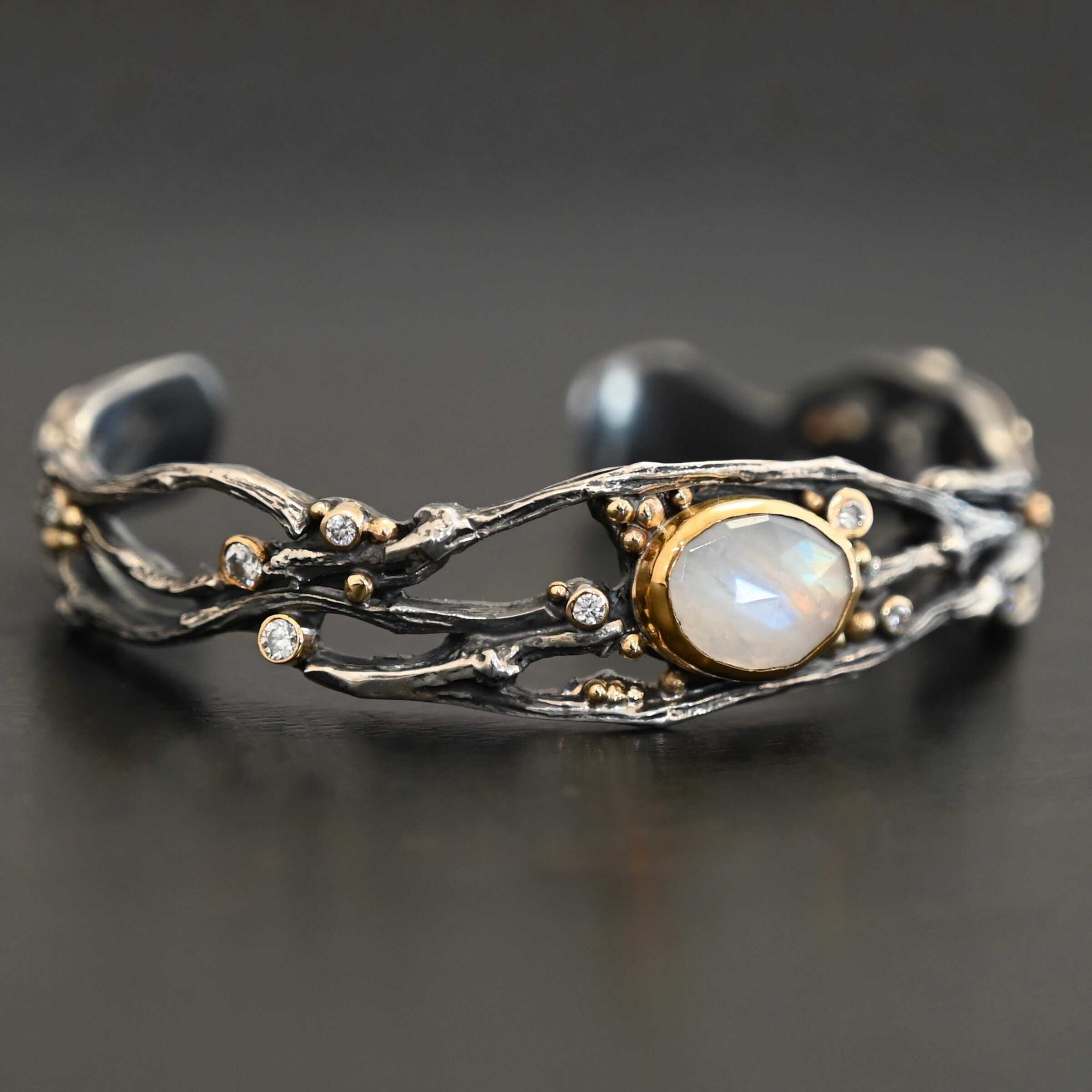 Night Flowers Cuff "Night Flowers" Cuff. There is a moonstone set in gold in the center of a cuff. The band of the cuff is multiple casted sterling silver twigs that intercept. There are small diamonds scattered around it, as well as gold ball detail.