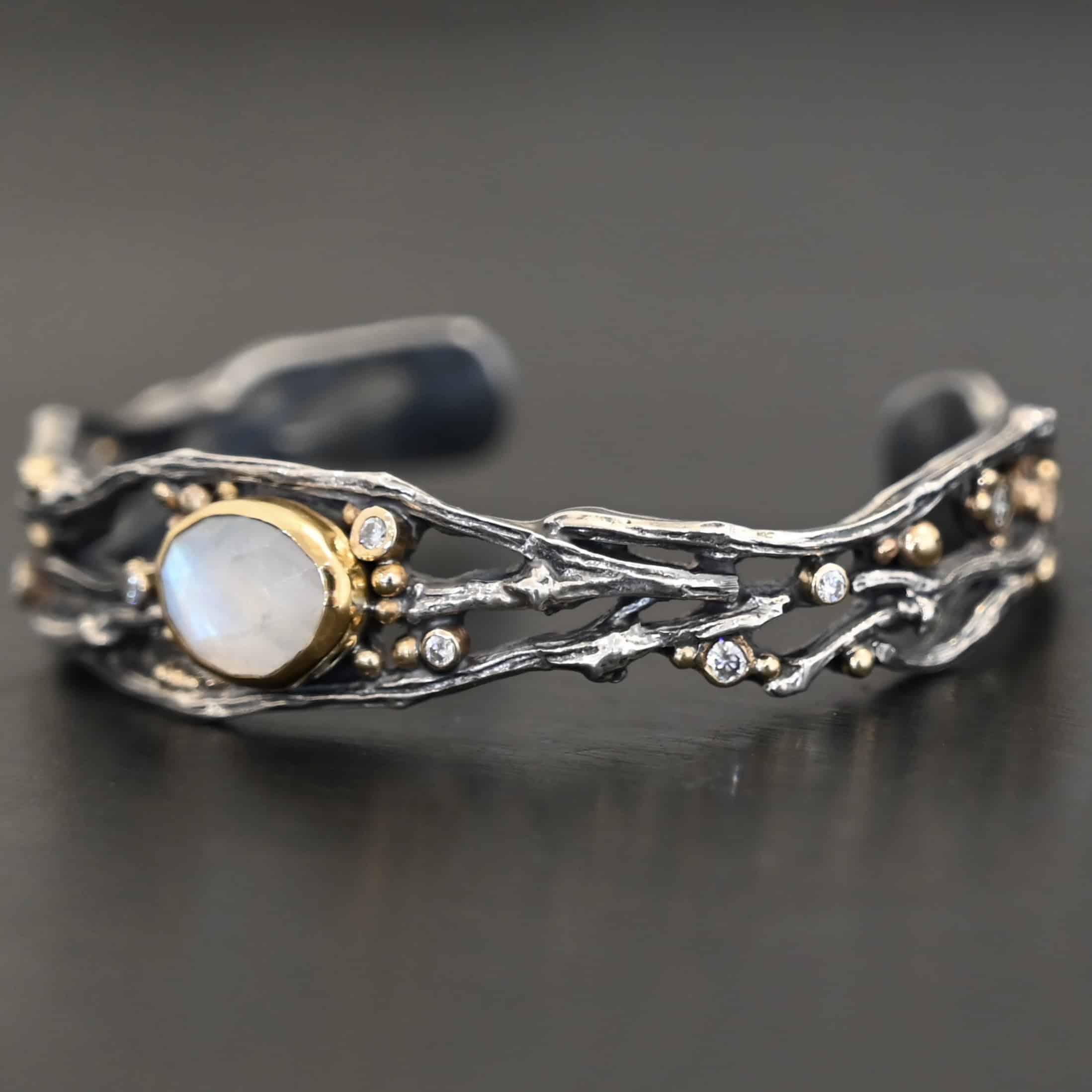 Night Flowers Cuff "Night Flowers" Cuff. There is a moonstone set in gold in the center of a cuff. The band of the cuff is multiple casted sterling silver twigs that intercept. There are small diamonds scattered around it, as well as gold ball detail.