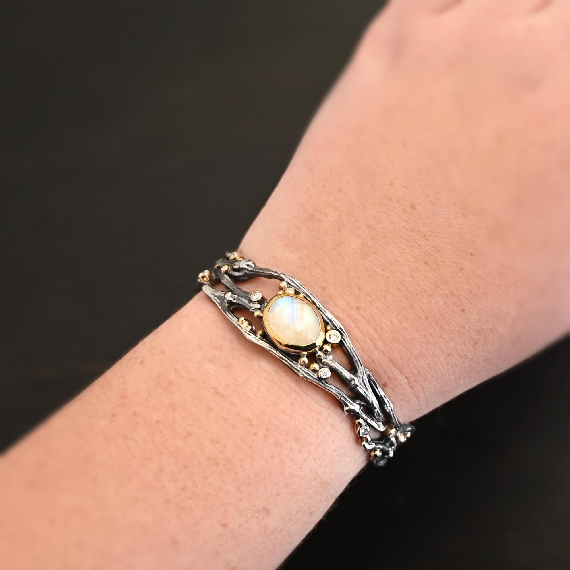 Night Flowers Cuff "Night Flowers" Cuff. There is a moonstone set in gold in the center of a cuff. The band of the cuff is multiple casted sterling silver twigs that intercept. There are small diamonds scattered around it, as well as gold ball detail. Cuff is shown up close by a model.