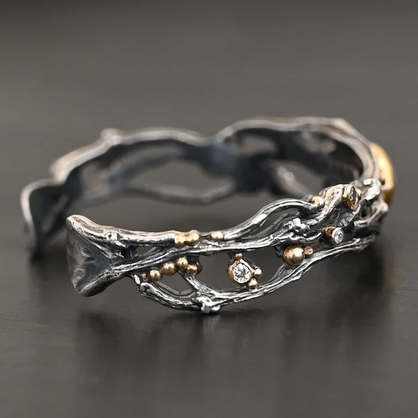 Night Flowers Cuff "Night Flowers" Cuff. There is a moonstone set in gold in the center of a cuff. The band of the cuff is multiple casted sterling silver twigs that intercept. There are small diamonds scattered around it, as well as gold ball detail. Cuff is shown up close by a model.