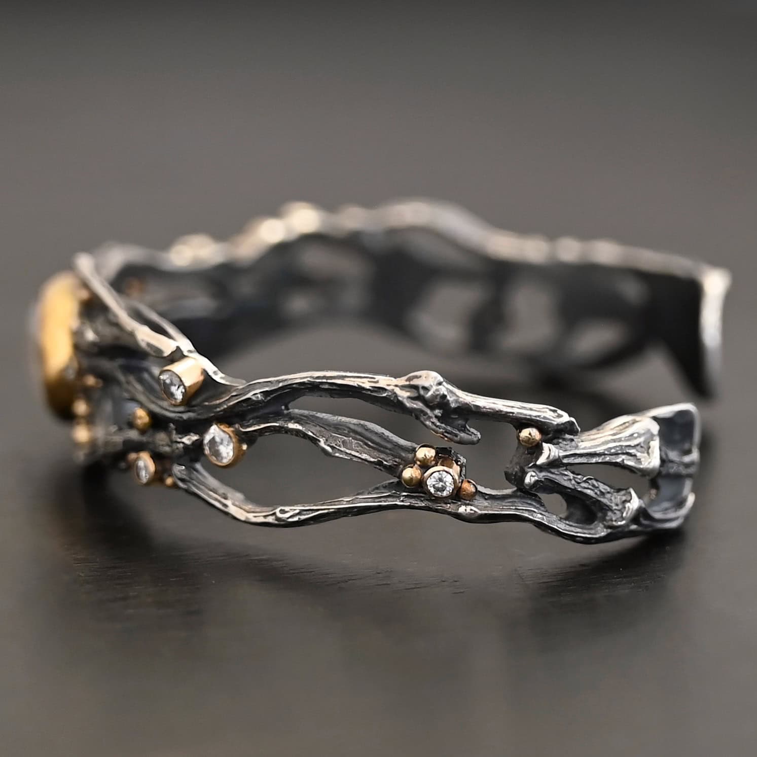 Night Flowers Cuff "Night Flowers" Cuff. There is a moonstone set in gold in the center of a cuff. The band of the cuff is multiple casted sterling silver twigs that intercept. There are small diamonds scattered around it, as well as gold ball detail. Cuff is shown up close by a model.