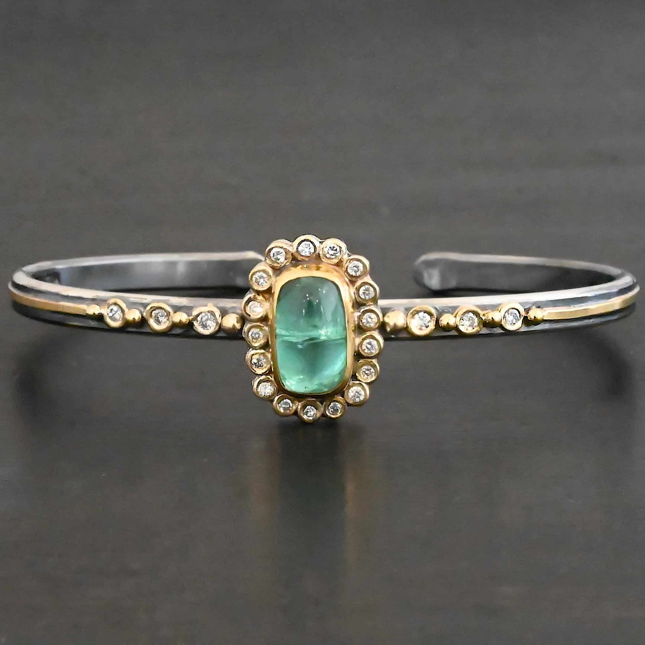 "Secret Of The Sea" Cuff. There is a rectangular, vertical tourmaline in the center, set in gold, with small diamonds surrounding the entire tourmaline. There are seventeen diamonds around the tourmaline. There are six more, three on each side of the cuff. There is gold ball detail between each diamond, and a stripe of gold that goes across the rest of the cuff.