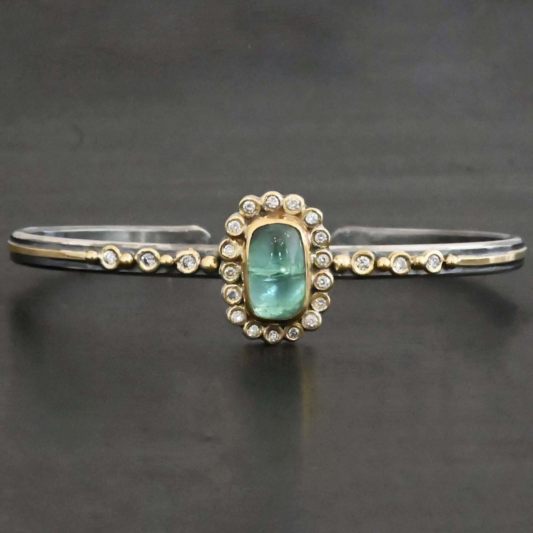 "Secret Of The Sea" Cuff. There is a rectangular, vertical tourmaline in the center, set in gold, with small diamonds surrounding the entire tourmaline. There are seventeen diamonds around the tourmaline. There are six more, three on each side of the cuff. There is gold ball detail between each diamond, and a stripe of gold that goes across the rest of the cuff.