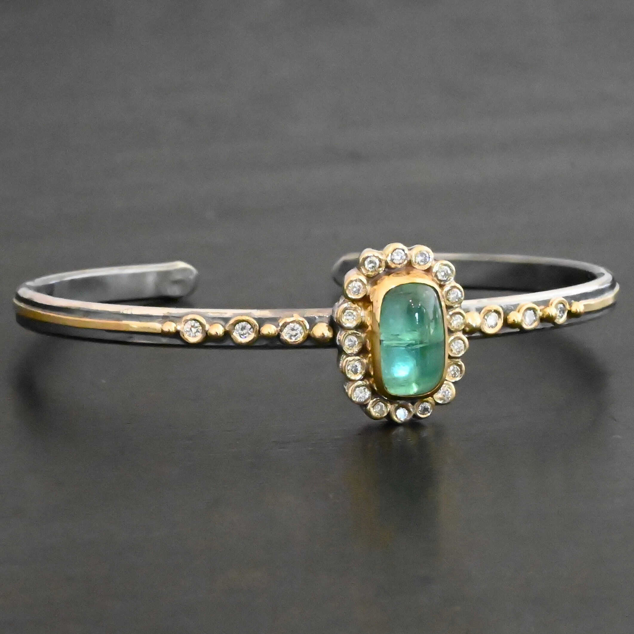 "Secret Of The Sea" Cuff. There is a rectangular, vertical tourmaline in the center, set in gold, with small diamonds surrounding the entire tourmaline. There are seventeen diamonds around the tourmaline. There are six more, three on each side of the cuff. There is gold ball detail between each diamond, and a stripe of gold that goes across the rest of the cuff.