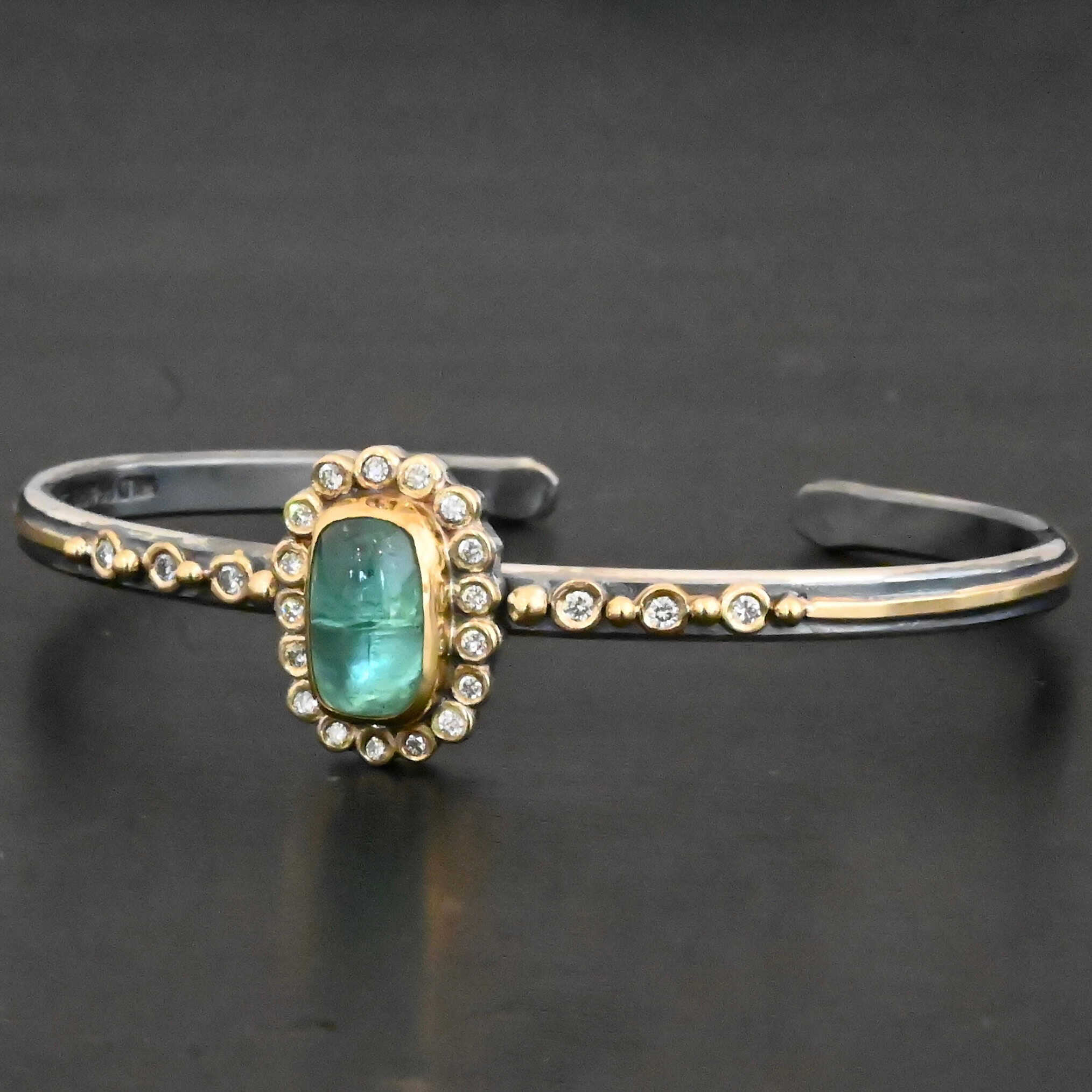 "Secret Of The Sea" Cuff. There is a rectangular, vertical tourmaline in the center, set in gold, with small diamonds surrounding the entire tourmaline. There are seventeen diamonds around the tourmaline. There are six more, three on each side of the cuff. There is gold ball detail between each diamond, and a stripe of gold that goes across the rest of the cuff.