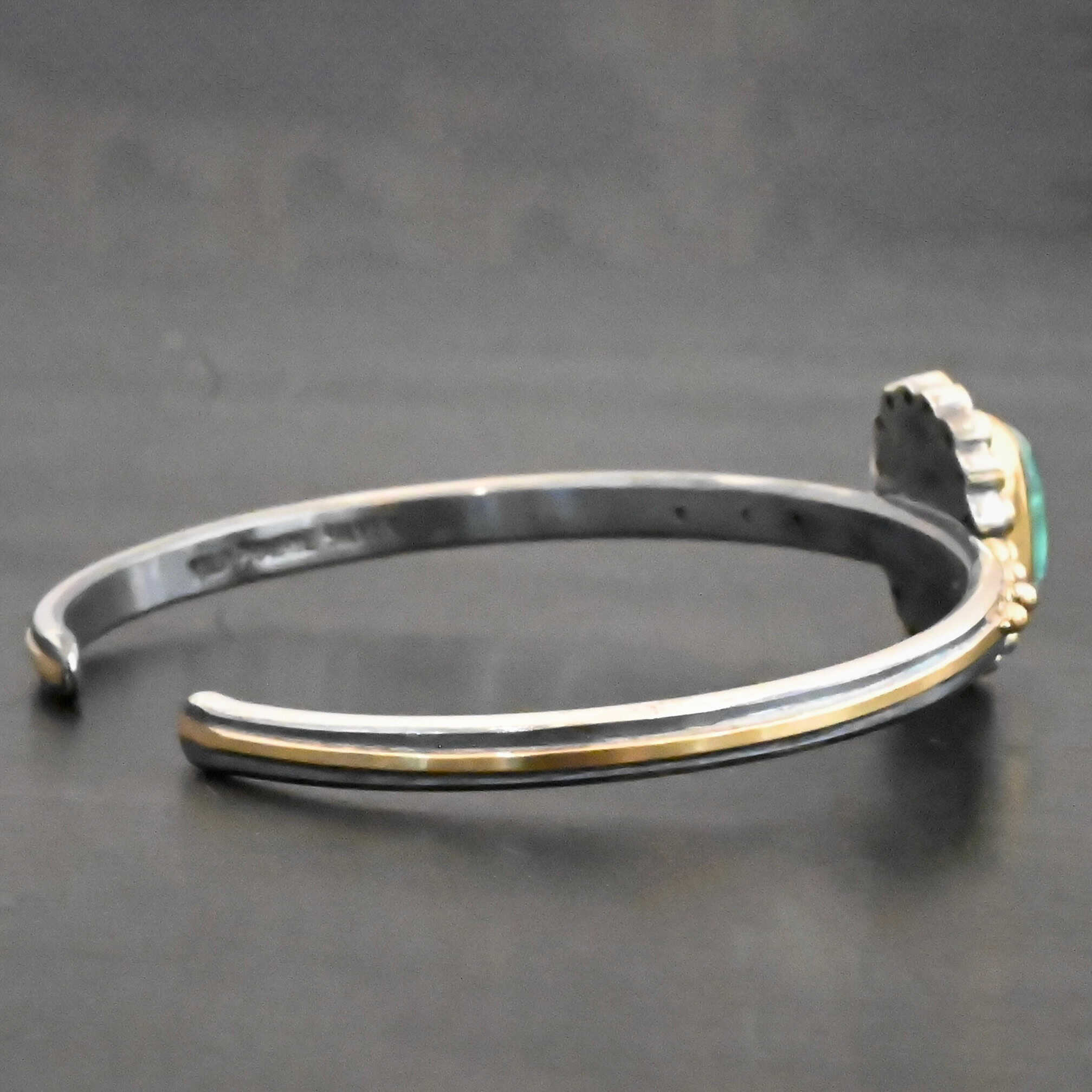 "Secret Of The Sea" Cuff. There is a rectangular, vertical tourmaline in the center, set in gold, with small diamonds surrounding the entire tourmaline. There are seventeen diamonds around the tourmaline. There are six more, three on each side of the cuff. There is gold ball detail between each diamond, and a stripe of gold that goes across the rest of the cuff.