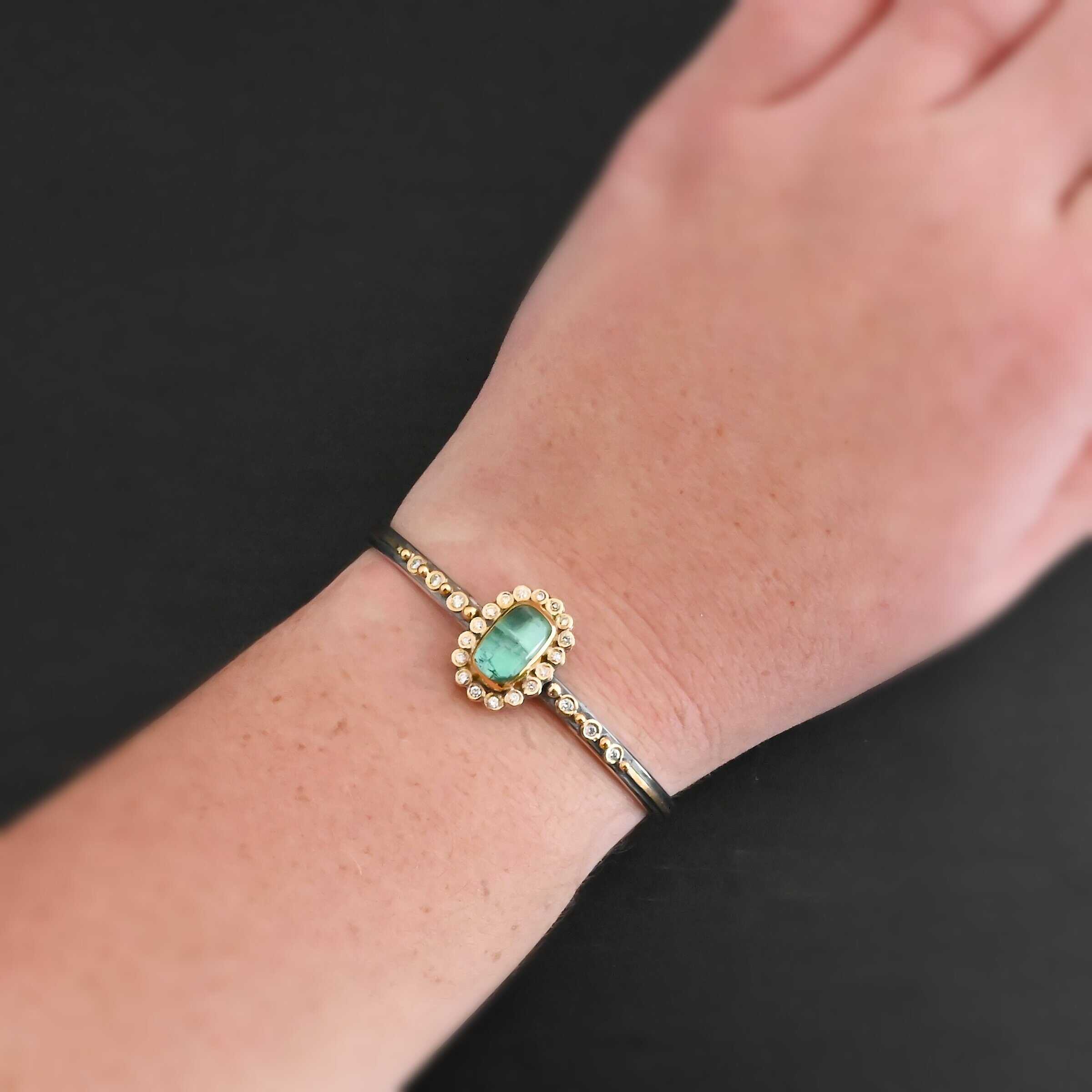 "Secret Of The Sea" Cuff. There is a rectangular, vertical tourmaline in the center, set in gold, with small diamonds surrounding the entire tourmaline. There are seventeen diamonds around the tourmaline. There are six more, three on each side of the cuff. There is gold ball detail between each diamond, and a stripe of gold that goes across the rest of the cuff. Cuff is shown up close by a model.