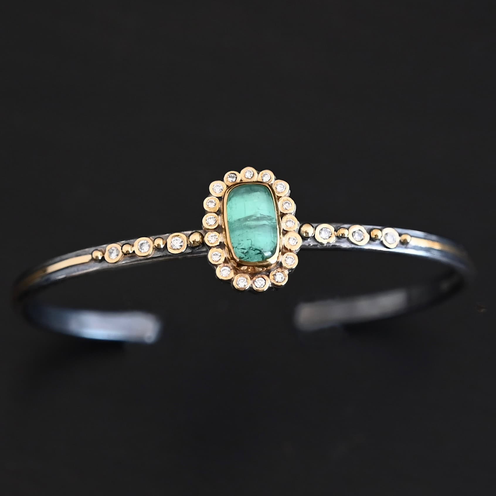 "Secret Of The Sea" Cuff. There is a rectangular, vertical tourmaline in the center, set in gold, with small diamonds surrounding the entire tourmaline. There are seventeen diamonds around the tourmaline. There are six more, three on each side of the cuff. There is gold ball detail between each diamond, and a stripe of gold that goes across the rest of the cuff.