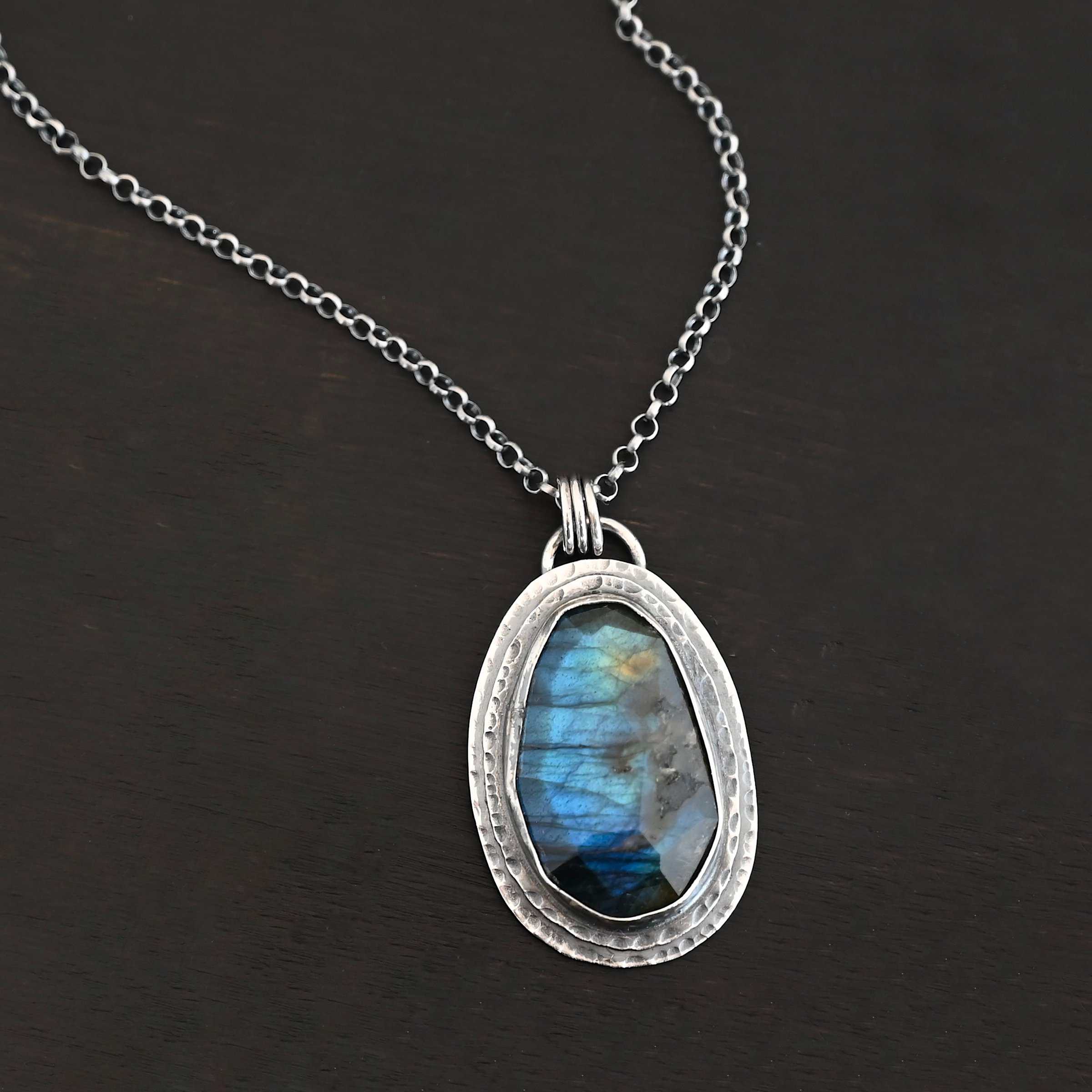 A New Day Necklace. There is a large, rose cut, vertical, oval labradorite in the center with two layers of sterling silver around it. There is a loop soldered to the back with three jump rings attached to it that the chain of the necklace goes through.