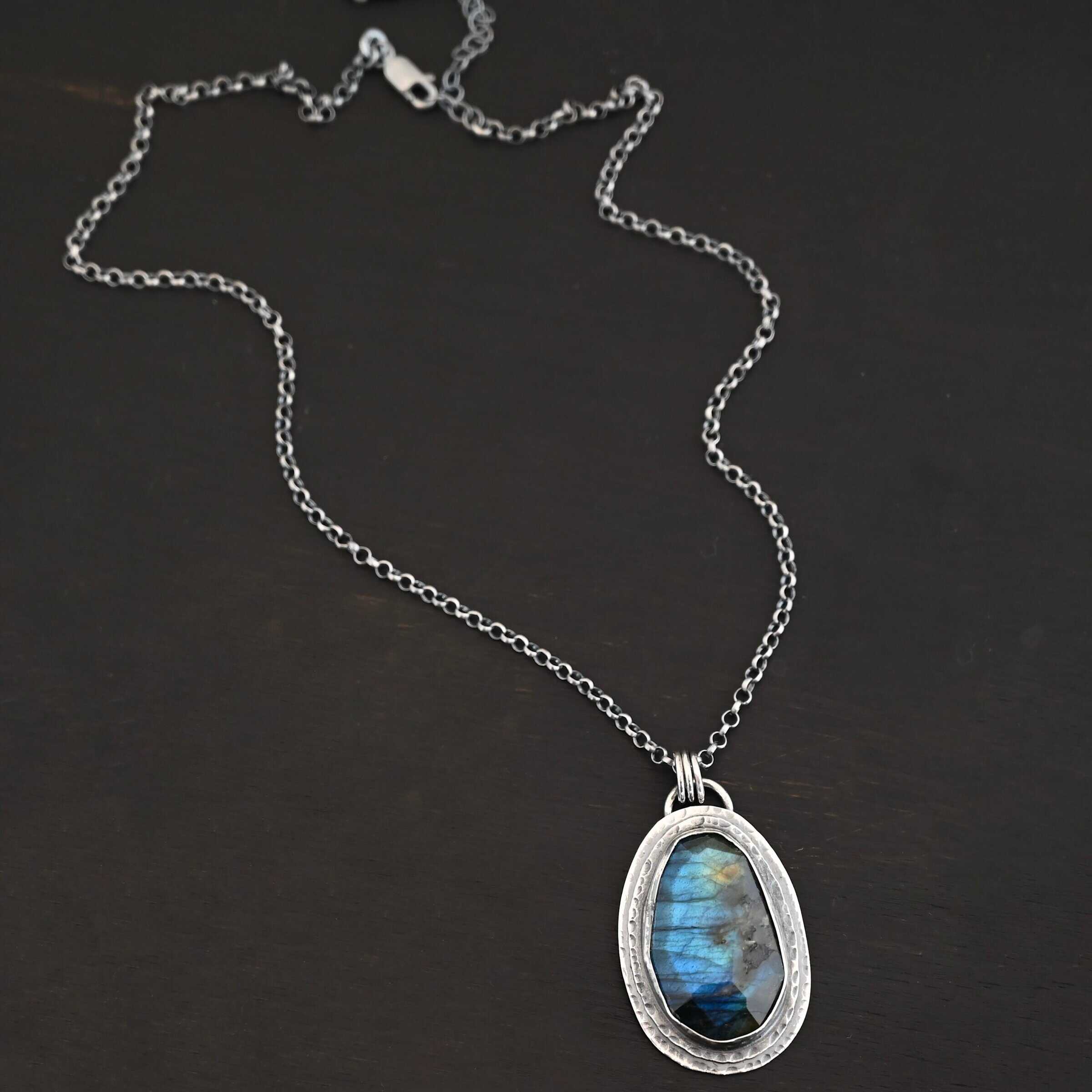 A New Day Necklace. There is a large, rose cut, vertical, oval labradorite in the center with two layers of sterling silver around it. There is a loop soldered to the back with three jump rings attached to it that the chain of the necklace goes through.