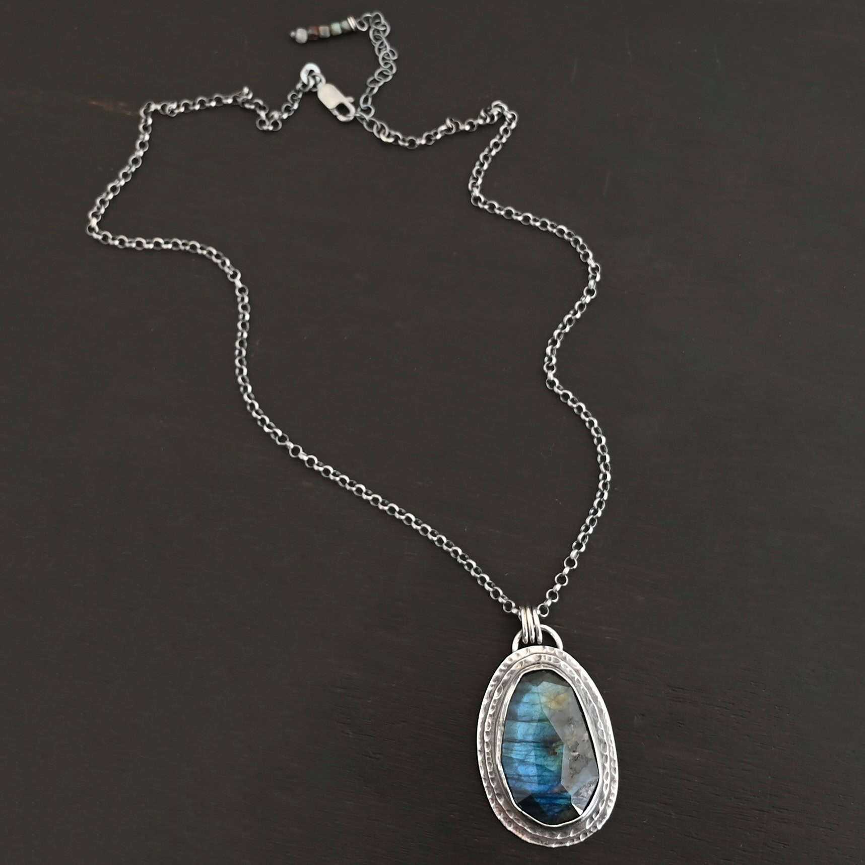 A New Day Necklace. There is a large, rose cut, vertical, oval labradorite in the center with two layers of sterling silver around it. There is a loop soldered to the back with three jump rings attached to it that the chain of the necklace goes through.