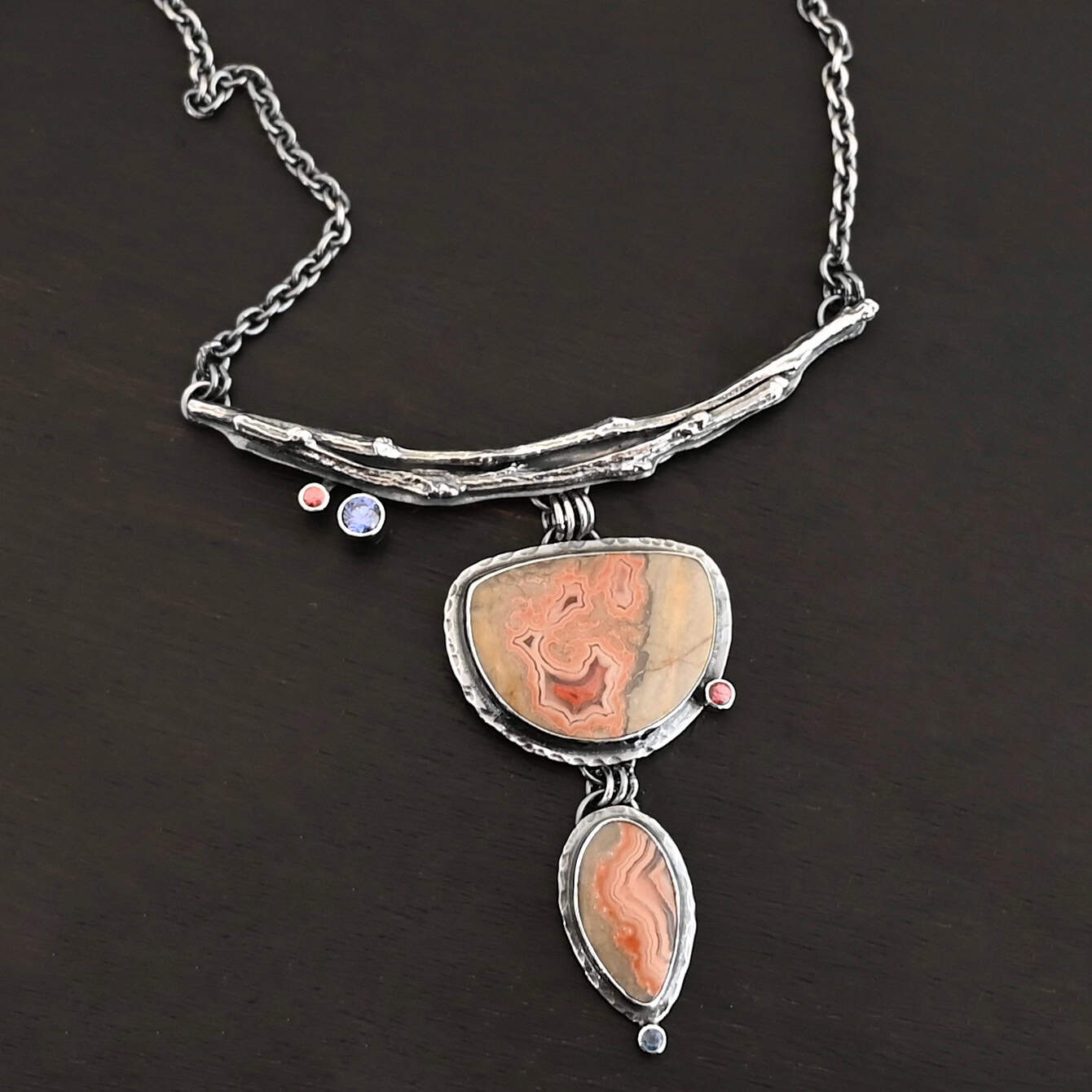 Dare To Be Necklace. There are two curved, casted sterling silver twigs from a branch at the top. On the left side bottom are two small stones. Hanging from the center is a semicircle ocean jasper with a small circular stone on the right side, and beneath that is an oval ocean jasper, vertical. On the bottom of that is a small circular stone.