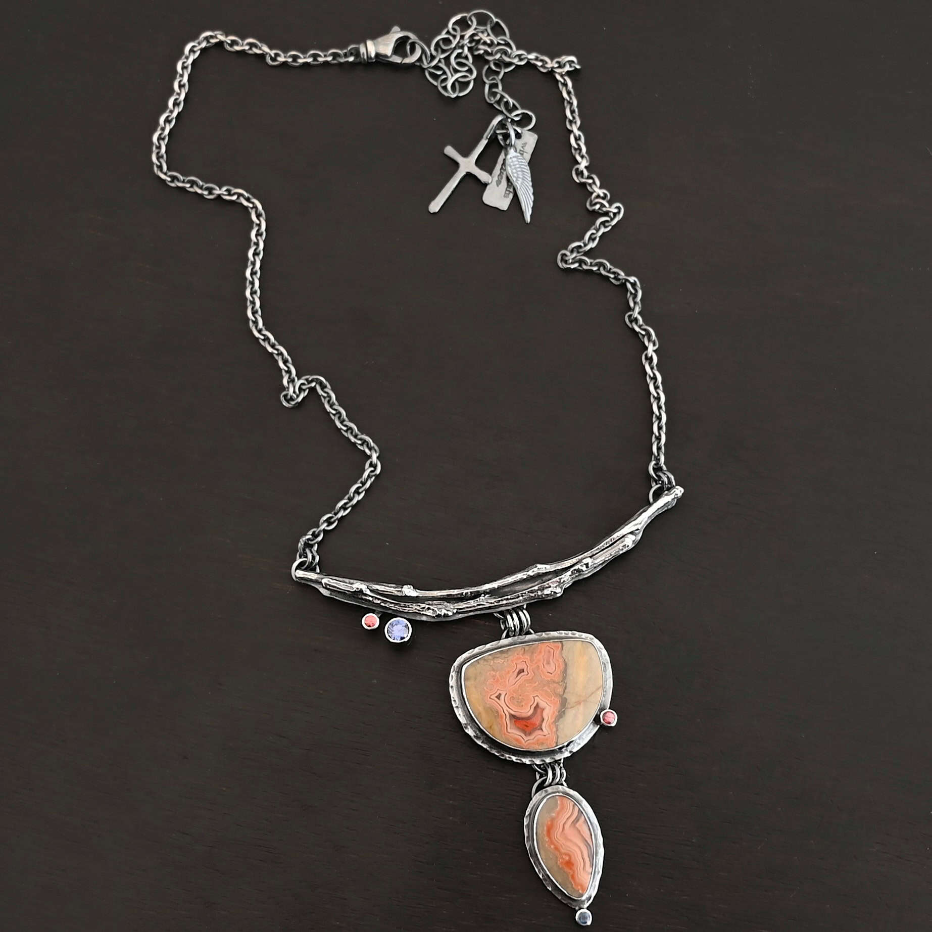 Dare To Be Necklace. There are two curved, casted sterling silver twigs from a branch at the top. On the left side bottom are two small stones. Hanging from the center is a semicircle ocean jasper with a small circular stone on the right side, and beneath that is an oval ocean jasper, vertical. On the bottom of that is a small circular stone.