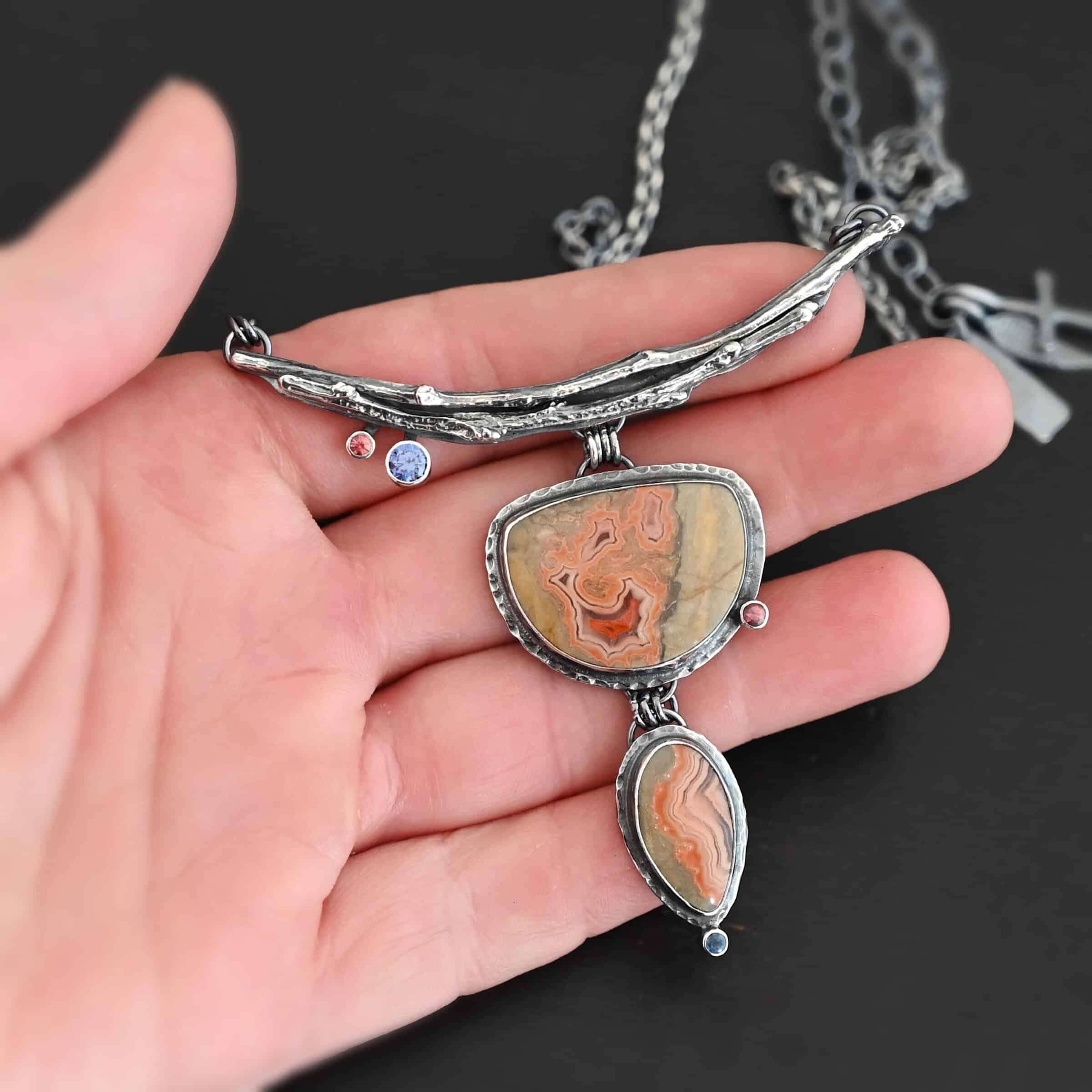 Dare To Be Necklace. There are two curved, casted sterling silver twigs from a branch at the top. On the left side bottom are two small stones. Hanging from the center is a semicircle ocean jasper with a small circular stone on the right side, and beneath that is an oval ocean jasper, vertical. On the bottom of that is a small circular stone. Necklace is held in the fingertips of a hand.