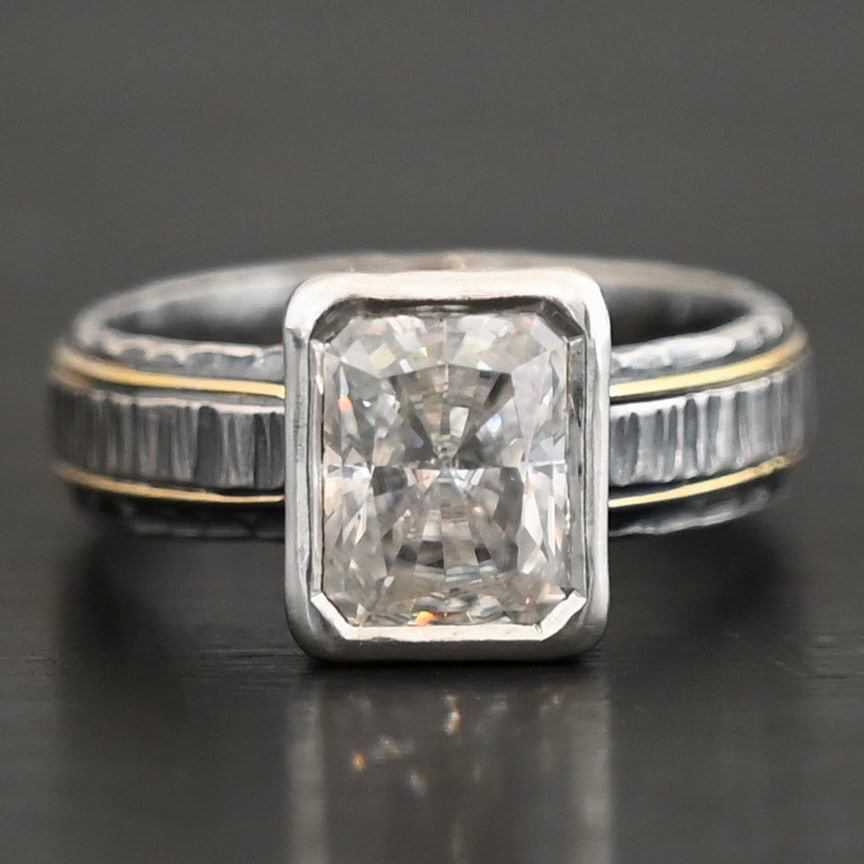 Photo of the "Love & Be Loved" Ring. There is a rectangular moissanite, set vertically in the center in sterling silver. The band of the ring is sterling silver as well, with a thick wrap of sterling silver around it that is raised and hammered with lines. There are two thin lines of gold that circle around the top and bottom of the ring. Ring is set against a dark background.