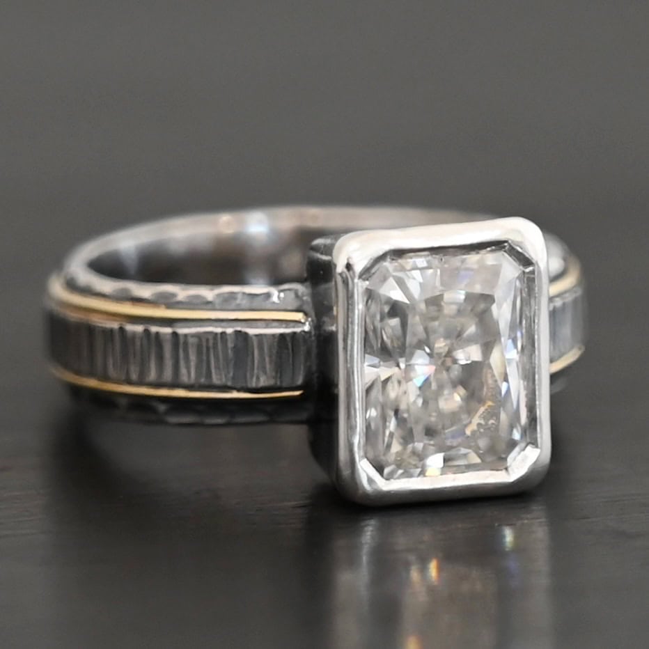 Photo of the "Love & Be Loved" Ring. There is a rectangular moissanite, set vertically in the center in sterling silver. The band of the ring is sterling silver as well, with a thick wrap of sterling silver around it that is raised and hammered with lines. There are two thin lines of gold that circle around the top and bottom of the ring. Ring is set against a dark background.