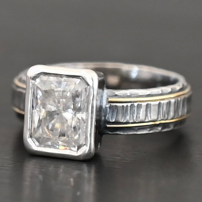 Photo of the "Love & Be Loved" Ring. There is a rectangular moissanite, set vertically in the center in sterling silver. The band of the ring is sterling silver as well, with a thick wrap of sterling silver around it that is raised and hammered with lines. There are two thin lines of gold that circle around the top and bottom of the ring. Ring is set against a dark background.