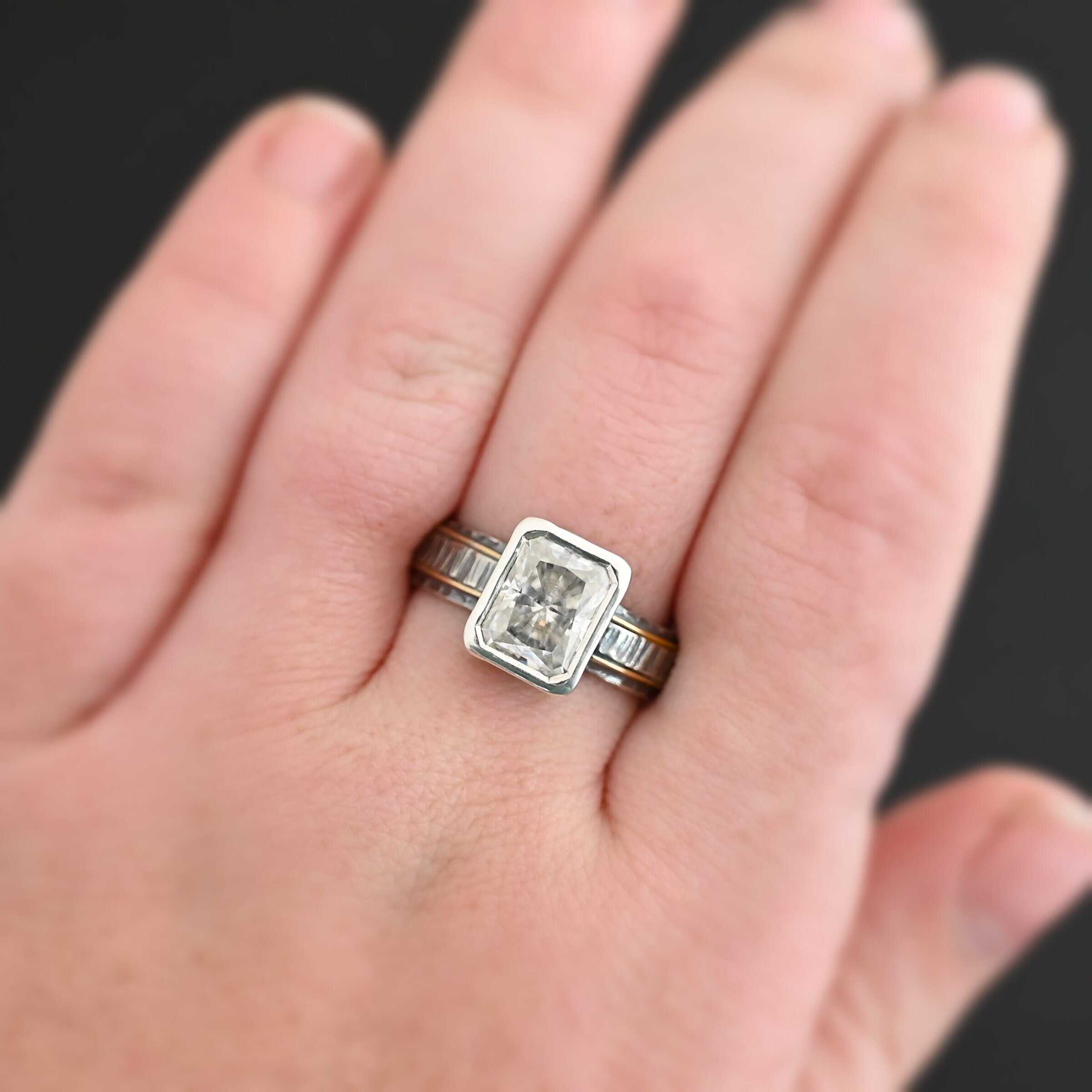 Photo of the "Love & Be Loved" Ring. There is a rectangular moissanite, set vertically in the center in sterling silver. The band of the ring is sterling silver as well, with a thick wrap of sterling silver around it that is raised and hammered with lines. There are two thin lines of gold that circle around the top and bottom of the ring. Ring is worn on a middle finger up close by a model to demonstrate size.