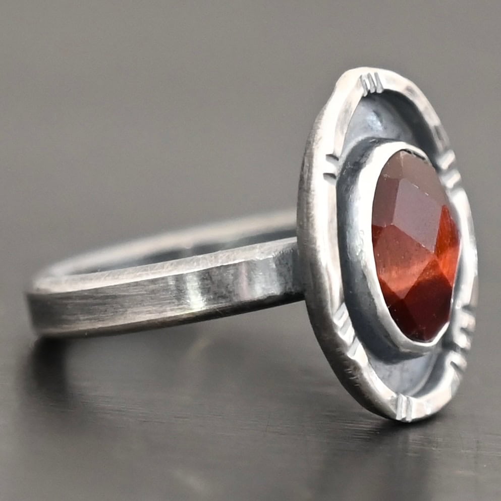 Ember Ring Photo of the "Ember" Ring. There is an oval, rose cut pietersite in the center. There is a ring of silver around it that is smooth and oxidized. There is another ring of silver around this that is lightly hammered with lines around it, and lightly oxidized.