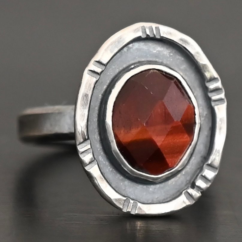 Ember Ring Photo of the "Ember" Ring. There is an oval, rose cut pietersite in the center. There is a ring of silver around it that is smooth and oxidized. There is another ring of silver around this that is lightly hammered with lines around it, and lightly oxidized.