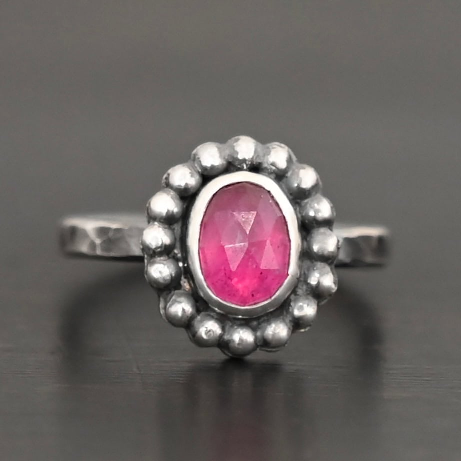 Emilia Ring Photo of the "Emilia" Ring. The ring band is smooth and oxidized silver. There is an oval ruby in the center with soldered balls of silver circling all the way around the ruby. Ring is sitting against a dark background.