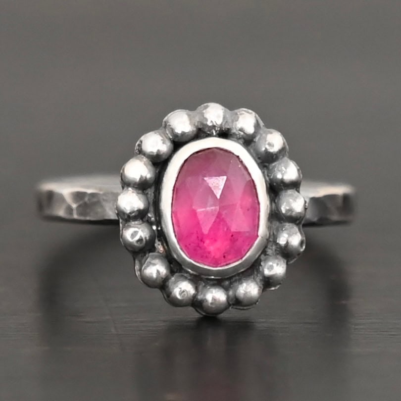 Emilia Ring Photo of the "Emilia" Ring. The ring band is smooth and oxidized silver. There is an oval ruby in the center with soldered balls of silver circling all the way around the ruby. Ring is sitting against a dark background.