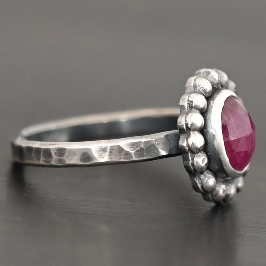 Emilia Ring Photo of the "Emilia" Ring. The ring band is smooth and oxidized silver. There is an oval ruby in the center with soldered balls of silver circling all the way around the ruby. Ring is sitting against a dark background.
