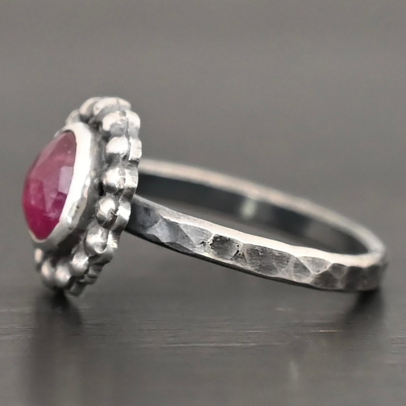 Emilia Ring Photo of the "Emilia" Ring. The ring band is smooth and oxidized silver. There is an oval ruby in the center with soldered balls of silver circling all the way around the ruby. Ring is sitting against a dark background.