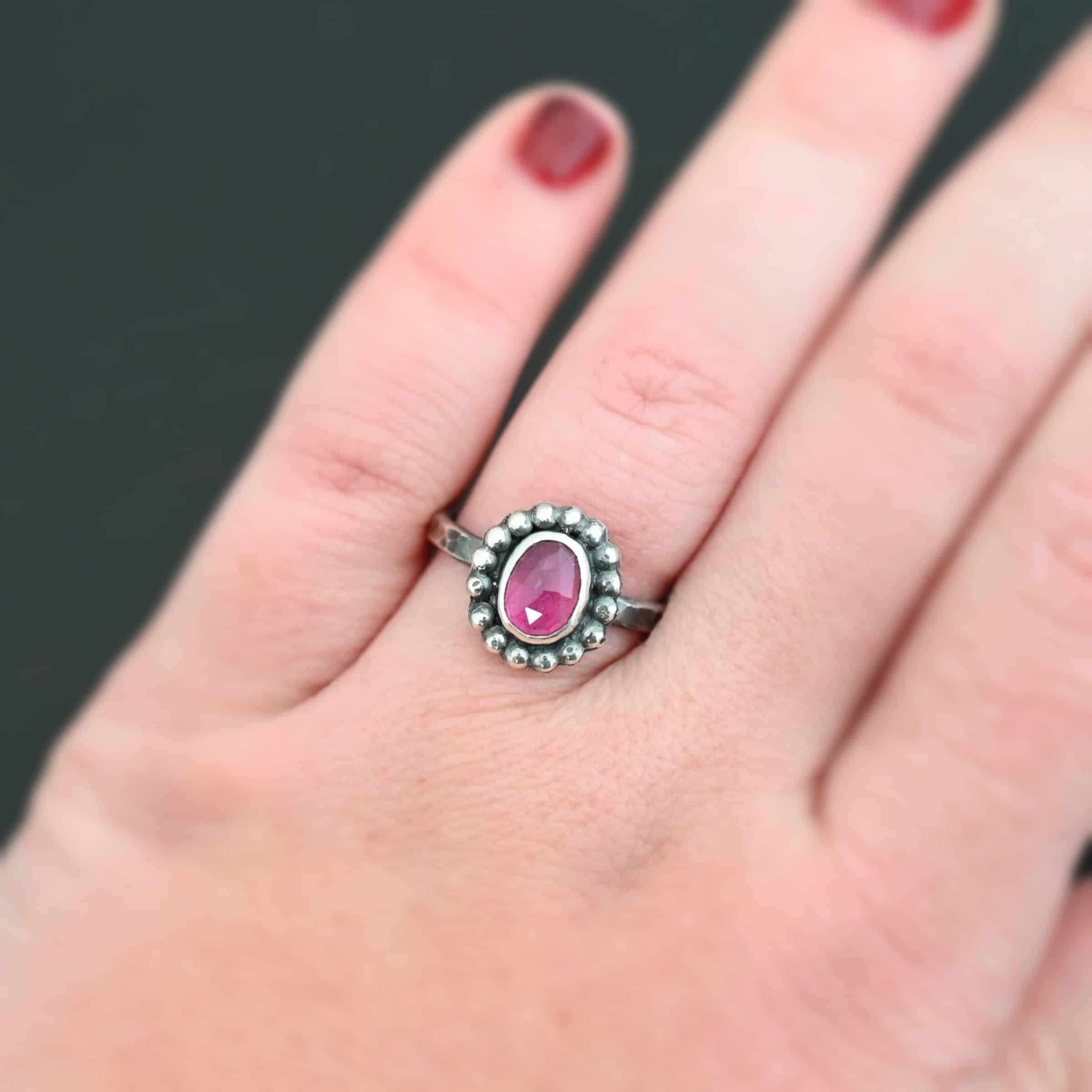 Emilia Ring Photo of the "Emilia" Ring. The ring band is smooth and oxidized silver. There is an oval ruby in the center with soldered balls of silver circling all the way around the ruby. Ring is worn by a model to demonstrate size.
