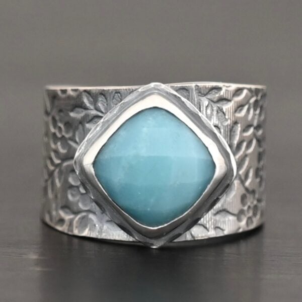 Photo of the "Naia" Ring. There is a medium sized, diamond shaped aquamarine in the center of the ring. The ring band is wide, oxidized lightly, sterling silver, with flower designs etched all the way around the entire band. Ring is sitting against a dark background.