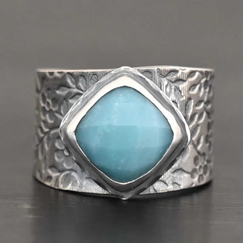 Naia Ring Photo of the "Naia" Ring. There is a medium sized, diamond shaped aquamarine in the center of the ring. The ring band is wide, oxidized lightly, sterling silver, with flower designs etched all the way around the entire band. Ring is sitting against a dark background.