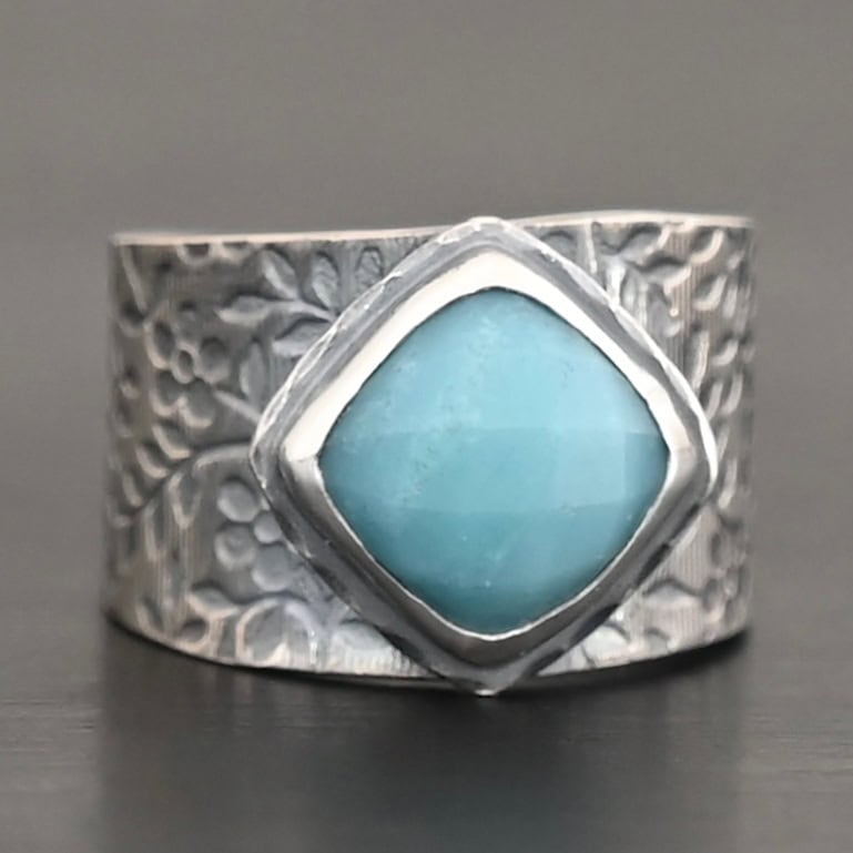 Naia Ring Photo of the "Naia" Ring. There is a medium sized, diamond shaped aquamarine in the center of the ring. The ring band is wide, oxidized lightly, sterling silver, with flower designs etched all the way around the entire band. Ring is sitting against a dark background.