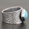 Photo of the "Naia" Ring. There is a medium sized, diamond shaped aquamarine in the center of the ring. The ring band is wide, oxidized lightly, sterling silver, with flower designs etched all the way around the entire band. Ring is sitting against a dark background.