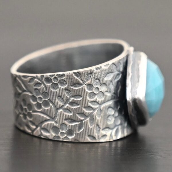 Photo of the "Naia" Ring. There is a medium sized, diamond shaped aquamarine in the center of the ring. The ring band is wide, oxidized lightly, sterling silver, with flower designs etched all the way around the entire band. Ring is sitting against a dark background.